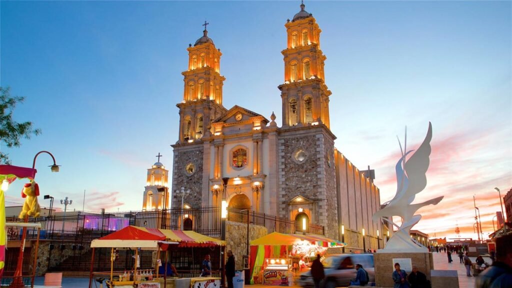 Catedral iluminada con puestos callejeros y escultura al atardecer.